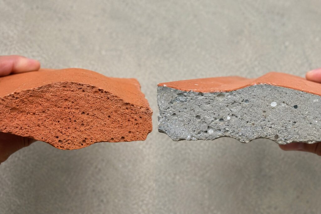 Clay vs concrete roof tiles broken edge comparison, solid terracotta clay core next to a concrete tile with gray cement core under an orange 